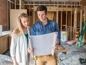 A man and woman are standing indoors, looking at a piece of paper. They both have smiles on their faces, and the background features a wall of a house. Both individuals are dressed in casual clothing.