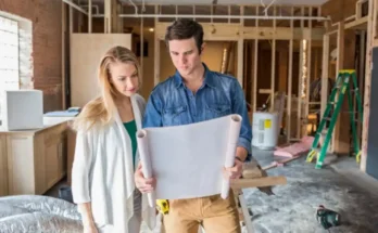 A man and woman are standing indoors, looking at a piece of paper. They both have smiles on their faces, and the background features a wall of a house. Both individuals are dressed in casual clothing.