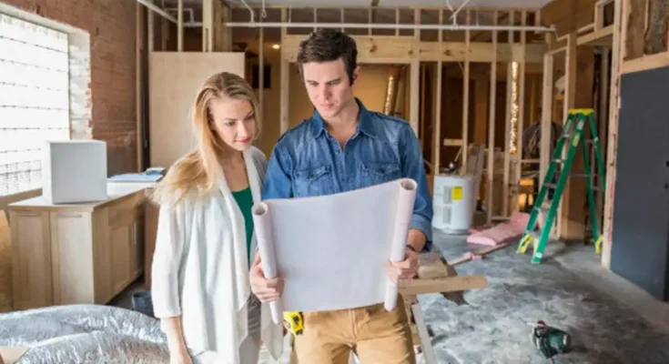 A man and woman are standing indoors, looking at a piece of paper. They both have smiles on their faces, and the background features a wall of a house. Both individuals are dressed in casual clothing.