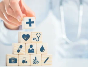 A person stacks wooden blocks with medical icons, such as a cross, heart, stethoscope, and syringe, symbolizing various aspects of healthcare.