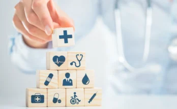 A person stacks wooden blocks with medical icons, such as a cross, heart, stethoscope, and syringe, symbolizing various aspects of healthcare.