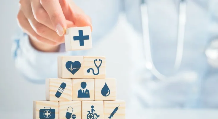 A person stacks wooden blocks with medical icons, such as a cross, heart, stethoscope, and syringe, symbolizing various aspects of healthcare.