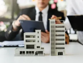 A model of a building on a table with a man dressed in professional attire using a tablet while sitting nearby in an indoor office environment.