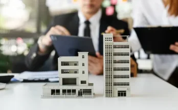 A model of a building on a table with a man dressed in professional attire using a tablet while sitting nearby in an indoor office environment.