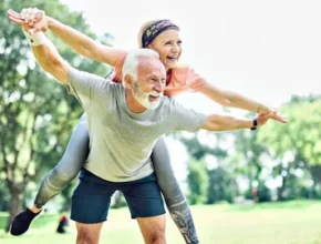 A woman enjoys a playful ride on an old man's back indicating fitness while both smiling, surrounded by greenery in a sunny park.