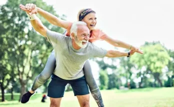 A woman enjoys a playful ride on an old man's back indicating fitness while both smiling, surrounded by greenery in a sunny park.