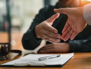 Two hands reaching for a handshake over a legal document on a wooden table, with a gavel in the foreground.