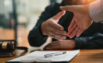 Two hands reaching for a handshake over a legal document on a wooden table, with a gavel in the foreground.