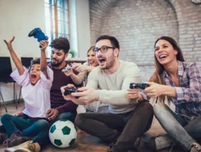 A group of friends sitting on the floor, enthusiastically playing video games with controllers, and a soccer ball beside them.