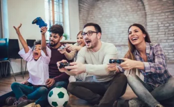 A group of friends sitting on the floor, enthusiastically playing video games with controllers, and a soccer ball beside them.