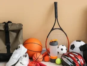 A collection of sports gear includes a backpack, various balls, a tennis racket, sneakers, a yoga mat, and protective gloves.