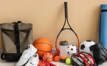 A collection of sports gear includes a backpack, various balls, a tennis racket, sneakers, a yoga mat, and protective gloves.