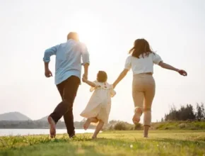 A family of three, a father, mother, and child, joyfully run barefoot together in a sunlit field by a river.