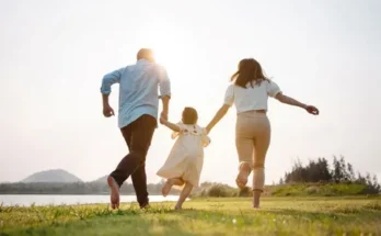 A family of three, a father, mother, and child, joyfully run barefoot together in a sunlit field by a river.