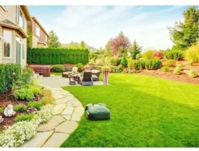 Lush green backyard with manicured lawn, stone path, cozy seating area, and vibrant flower beds under a bright blue sky.