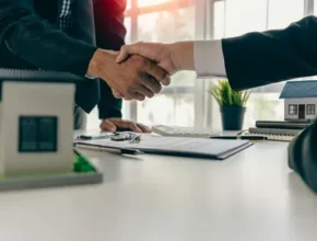 Business professionals shaking hands over a contract, with miniature houses on the table, symbolizing a real estate transaction.