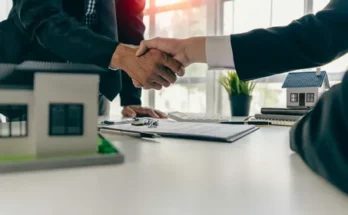 Business professionals shaking hands over a contract, with miniature houses on the table, symbolizing a real estate transaction.