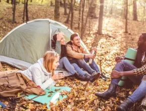 A group of friends relaxes by a tent in a sunlit forest, surrounded by autumn leaves, enjoying drinks and each other's company.