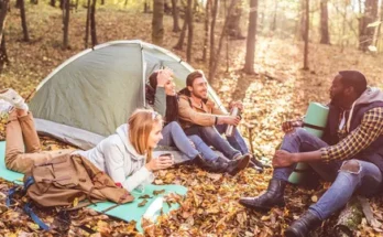 A group of friends relaxes by a tent in a sunlit forest, surrounded by autumn leaves, enjoying drinks and each other's company.