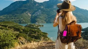 A woman in a hat and backpack captures a scenic view of mountains and a lake, enjoying nature on a sunny day.