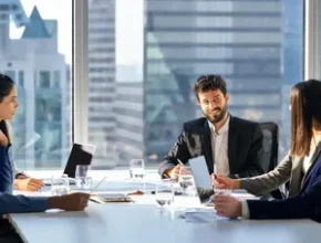 A diverse group of professionals in business attire engages in a meeting around a modern conference table, with a city skyline in the background.