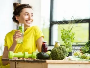 A woman in a bright yellow shirt enjoys a drink surrounded by fresh vegetables, juice bottles, and workout weights on a wooden table.
