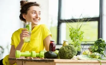 A woman in a bright yellow shirt enjoys a drink surrounded by fresh vegetables, juice bottles, and workout weights on a wooden table.