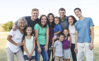 A diverse group of family members poses together outdoors, showcasing smiles and a range of casual summer outfits in a sunny setting.
