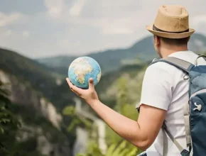 A traveler holding a globe stands atop a mountain, gazing at a scenic landscape with rolling hills and a blue sky.