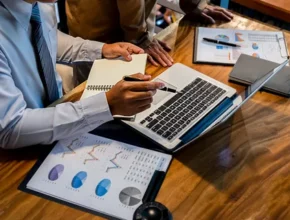 A businessman in a white shirt and tie analyzes data on a laptop, with charts and notes on a wooden table.