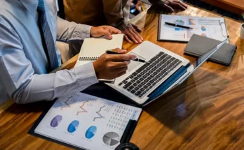 A businessman in a white shirt and tie analyzes data on a laptop, with charts and notes on a wooden table.