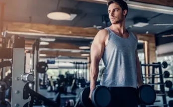 A muscular individual in a gray tank top lifts dumbbells in a well-equipped gym, showcasing strength and focus.