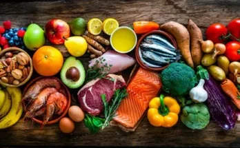 A vibrant assortment of fresh fruits, vegetables, seafood, meat, and nuts arranged on a wooden surface, showcasing healthy food choices.