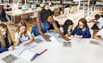 A teacher guides students at a library table, as they collaborate on worksheets surrounded by computers and bookshelves.