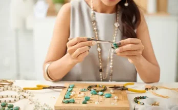 A woman carefully assembles jewelry with turquoise stones and tools on a workspace filled with various beads and materials.
