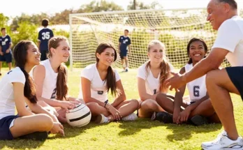 A group of female soccer players sit on the grass, listening intently to their coach, with a goal net visible in the background.