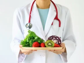 A healthcare professional in a white coat holds a wooden plate filled with fresh fruits and vegetables, symbolizing healthy eating.