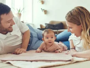 A man and woman lying on the floor together, with a baby resting between them. The setting is indoors, and they appear comfortable and relaxed.