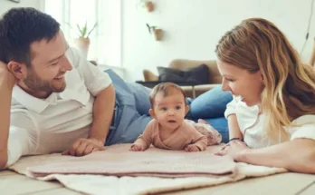 A man and woman lying on the floor together, with a baby resting between them. The setting is indoors, and they appear comfortable and relaxed.