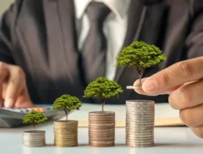 A businessman in a suit places a tree on a stack of coins, symbolizing financial growth and investment in sustainable practices.