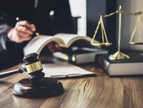A judge's gavel in the foreground with scales of justice and a lawyer studying legal texts in the background on a wooden table.