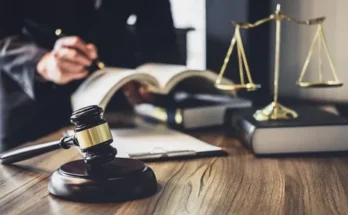 A judge's gavel in the foreground with scales of justice and a lawyer studying legal texts in the background on a wooden table.