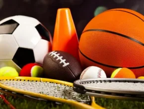A collection of various sports equipment, including soccer and basketballs, tennis racquets, and a football, on a grassy surface.