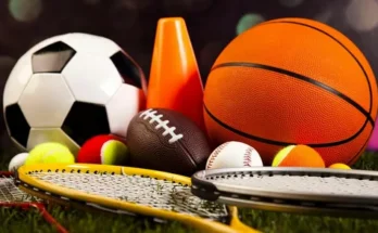 A collection of various sports equipment, including soccer and basketballs, tennis racquets, and a football, on a grassy surface.