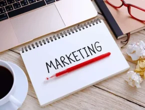 A notebook with "MARKETING" written in bold, a red pen, a coffee cup, glasses, and crumpled paper on a wooden desk.