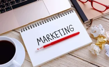 A notebook with "MARKETING" written in bold, a red pen, a coffee cup, glasses, and crumpled paper on a wooden desk.