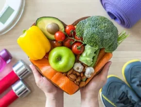 Hands holding a heart-shaped bowl filled with colorful fruits and vegetables, surrounded by workout equipment and a scale.