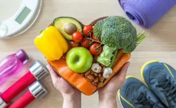 Hands holding a heart-shaped bowl filled with colorful fruits and vegetables, surrounded by workout equipment and a scale.