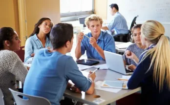A group of diverse students collaborate around a table, discussing ideas with laptops, tablets, and papers in a bright classroom.