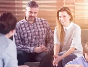 A family sits together in a cozy room with blinds, engaged in a happy discussion with another person about parenting.